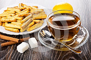 Bread sticks with poppy in plate, lemon, cinnamon sticks, cup of tea, spoon on saucer, sugar on wooden table