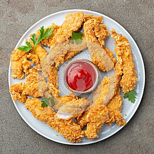 Breaded chicken strips with tomato ketchup on a white plate. Fast food on dark brown background. Top view