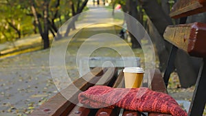 Break in autumn park, empty bench with knitted top and cup of coffee on walkway