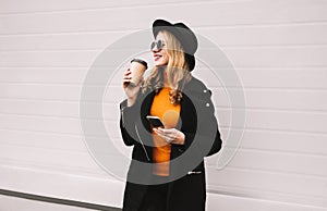 Break time! Fashion smiling woman enjoying coffee holds smartphone in city on gray