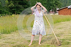 Break. Tired woman corrects headdress. A wooden rake is lean on her shoulder. Haymaking season (concept village life)