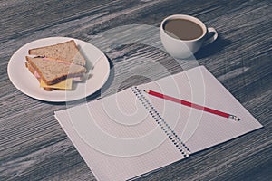 Break at work. Open copybook with a pencil, ham and cheese sandwich on a white plate, cup of hot tea on wooden background. Vintage