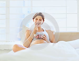 Breakfast in bed, eating and pregnant woman with strawberry bowl in a house, happy or hungry. Fruit, smile and female