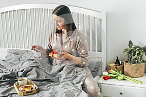 Breakfast in bed on Mother`s day. Young woman in pajamas holding gift box, reading card from family