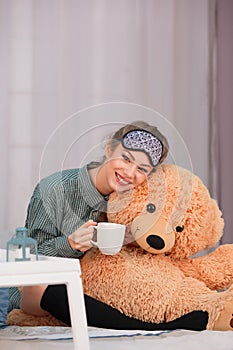 Breakfast in bed. Young woman smiles, drinks coffee tea and hugs big toy bear