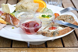 Breakfast with bread, butter and jam, also various cold cuts and fruits on a white plate on a wooden outdoor table, selected focus