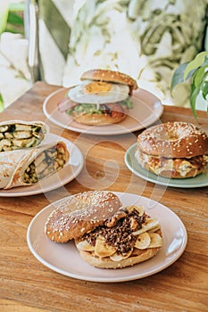 A breakfast burrito and bagel burger on a divided plate with salsa verde, sour cream, vegan, and chicken on a wooden table