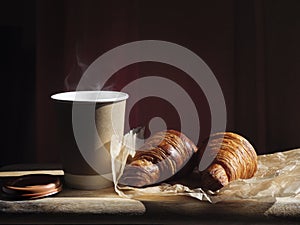 Breakfast with coffee cup and two croissants on a wooden table in sunlight.