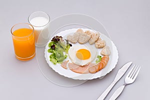 Breakfast consists of fried egg, ham, sausage, milk, orange, whole wheat bread shaped like a heart, fresh vegetables, put plates