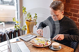Muž jesť. nôž a vidlica v ruke. z jesť na. prenosný počítač a raňajky na stôl. . americký štýl 