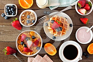 Breakfast food table scene. Fruits, cereal, waffles, yogurt and coffee. Top view over wood.