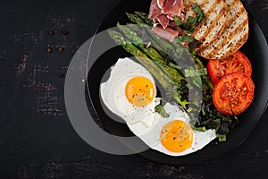 Breakfast. Fried egg, bread toast, green asparagus, tomatoes and jamon on black plate. Top view, above