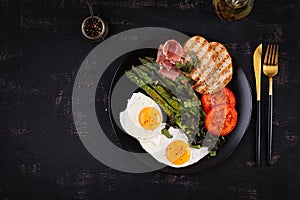 Breakfast. Fried egg, bread toast, green asparagus, tomatoes and jamon on black plate. Top view, above