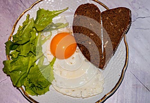 Breakfast with fried egg, slices bread with heard shape and greenery on white plate