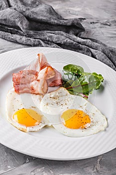 Breakfast, fried eggs, bacon, prosciutto, fresh salad on plate on grey table surface. Healthy food