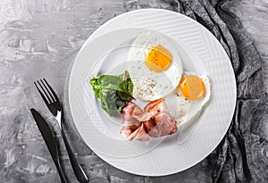Breakfast, fried eggs, bacon, prosciutto, fresh salad on plate on grey table surface. Healthy food, top view, flat lay