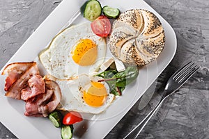 Breakfast, fried eggs, bun, bacon, prosciutto, fresh salad on plate on grey table surface. Healthy food
