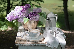 Breakfast in the garden: eclairs, cup of coffee, coffee pot, lilac flowers in a basket