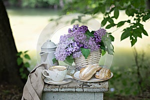 Breakfast in the garden: eclairs, cup of coffee, coffee pot, lilac flowers in a basket