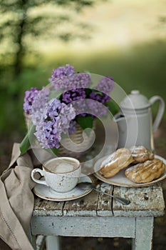 Breakfast in the garden: eclairs, cup of coffee, coffee pot, lilac flowers in a basket
