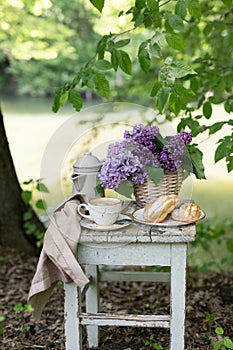 Breakfast in the garden: eclairs, cup of coffee, coffee pot, lilac flowers in a basket