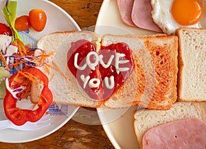 Breakfast have fried egg ham bread and salad. On the bread have heart drawing and text love you is made by ketchup and mayo.