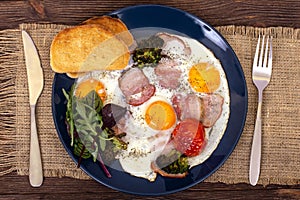 Breakfast or lunch with fried eggs, bread toast, arugula leaves, tomato, pork ham or bacon and broccoli on a dark blue plate