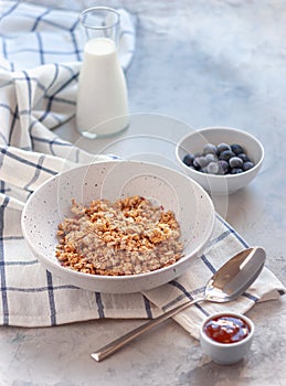 Breakfast, milk in a jug, muesli in a deep dish. Next to blueberries and jam on a blue-gray concrete background