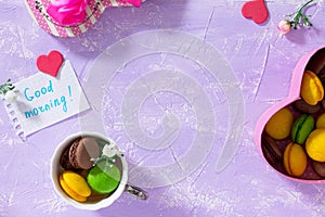 Breakfast, Mother`s Day, birthday background. Mug cakes Macaron, leaf notebook inscription good morning on purple table top.