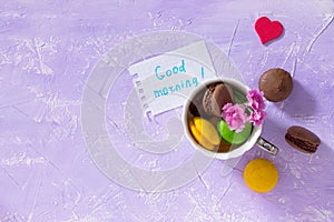 Breakfast, Mother`s Day, birthday background. Mug cakes Macaron, leaf notebook inscription good morning on purple table top.