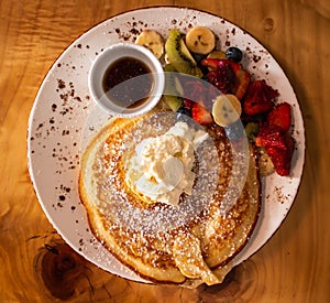 Breakfast pancake, maple syrup, cream, fresh fruit served on white plate atop wooden table