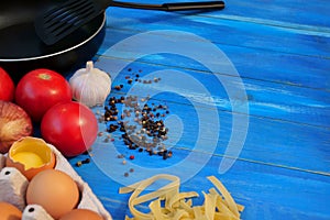 Breakfast Set. Frying pan, tomatoes, raw eggs, onions, garlic, spices, dry pasta. Selective focus. Copy space. On blue wooden