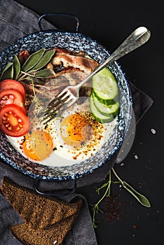 Breakfast set. Pan of fried eggs with bacon, fresh tomato, cucumber, sage and bread on dark serving board over black