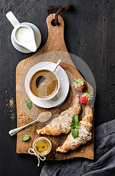 Breakfast set. Two freshly baked croissants with strawberries, honey, cup of black coffee, pitcher and spoon on rustic