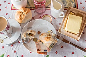 breakfast table with coffee sugar milk and butter and roll covered with euro coins and banknotes on plate, Germany