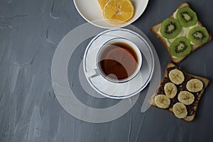 breakfast, tea with lemon and sandwiches, bread with chocolate, banana and kiwi slices on a gray background with space for text