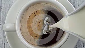 Breakfast time - adding milk to black coffee with foam. Pouring milk into coffee cup. Vintage white wooden background