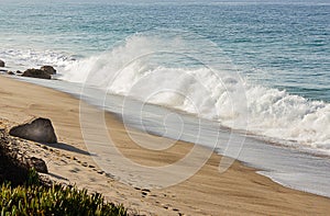 Breaking wave with backspray on a sandy beach with boulders and iceplant, with an ocean expanse horizon