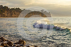 Breaking wave with foam and backwash curving across bay to penensulla and distant beach with rocky shoreline, at dawn