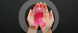 Breast cancer awareness symbol pink ribbon in woman hands on black background. Breast cancer awareness campaign banner