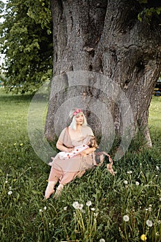 Breast feeding: Young hippie mother breastfeeds her baby girl child in city park sitting under a tree on a green grass