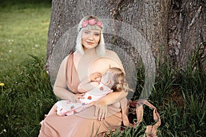 Breast feeding: Young hippie mother breastfeeds her baby girl child in city park sitting under a tree on a green grass