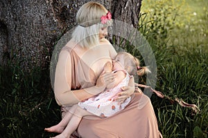 Breast feeding: Young hippie mother breastfeeds her baby girl child in city park sitting under a tree on a green grass