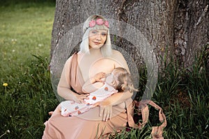 Breast feeding: Young hippie mother breastfeeds her baby girl child in city park sitting under a tree on a green grass