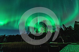 Breathtaking strong green lights of dancing Aurora over Northern forest, lake, small boat, bridge. Stocksjo lake, Northern Sweden