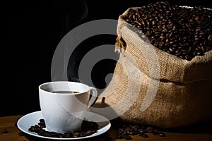 Brewed coffee in a white cup with steam or vapor and coffee beans in a burlap hessian sack and beans scattered on a wooden surface