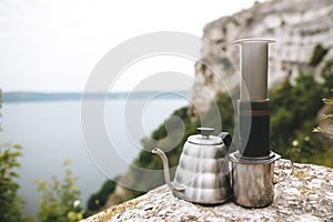 Brewing alternative coffee at camping. Steel kettle and aeropress on metal mug on cliff at lake, making hot drink at picnic