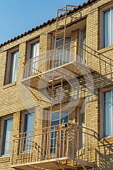 Brick building facade with brown fire escape ladders and platforms with large windows and blue sky background