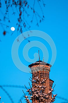 A brick minaret with a full moon in the sky.Double Minaret Madrasa ,Turkish: Cifte Minareli Medrese
