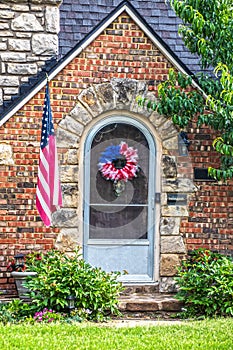 Brick and rock cottage with arched front door surrounded by strones with mounted American flag and red white and blue wreath on